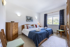 A hotel room with two beds, a wooden dresser, a desk, a chair, and a window with curtains. - CoolHouses Algarve Luz | Blue Ocean View T2 (Luz)