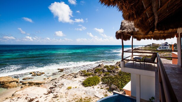A coastal view with a thatched roof balcony overlooking the sea, rocky shoreline, and distant buildings. - Casa Nido del Gallo Solar Ocean Front Deluxe Home 3 bedrooms, 5 bathrooms (Isla Mujeres)