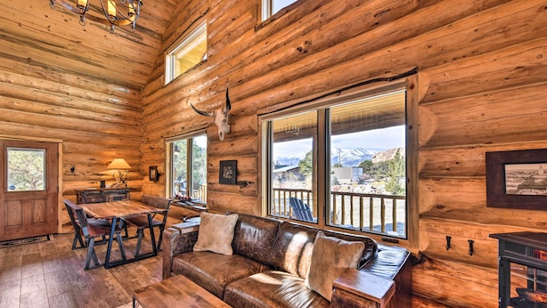 A log cabin interior with a wooden dining table, leather sofa, and large windows offering mountain views. - Riverfront Retreat w/ Hot Tub: Fish & Hike! (Buena Vista)