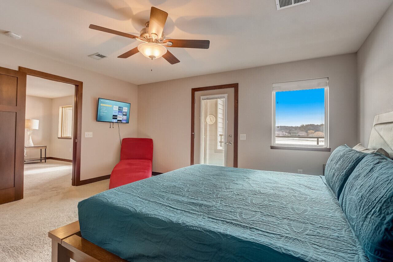 A bedroom with a bed, a ceiling fan, a TV, a red chair, and a window with a view.