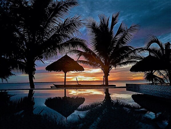 A serene sunset view with palm trees and a thatched umbrella by a reflective pool. - Asante Sana Beach Cottages - Beachfront Cottage  (Fukuchani)