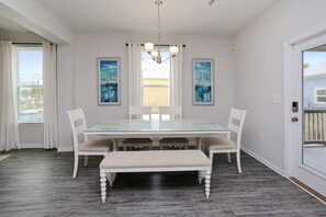 A dining room with a glass table, white chairs, a bench, and two paintings on the walls. - WindMark Beach, Community Pool, Playground, Short Walk to Beach ~ The Sandpiper (St. Joe Beach)