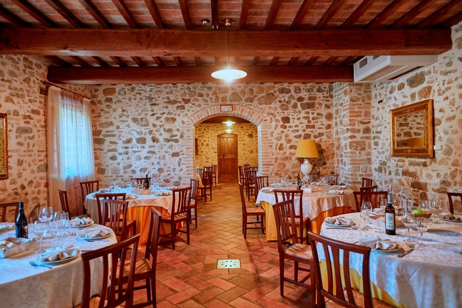 A restaurant with stone walls and wooden beams, tables set with white tablecloths and chairs, and a chandelier hanging from the ceiling.