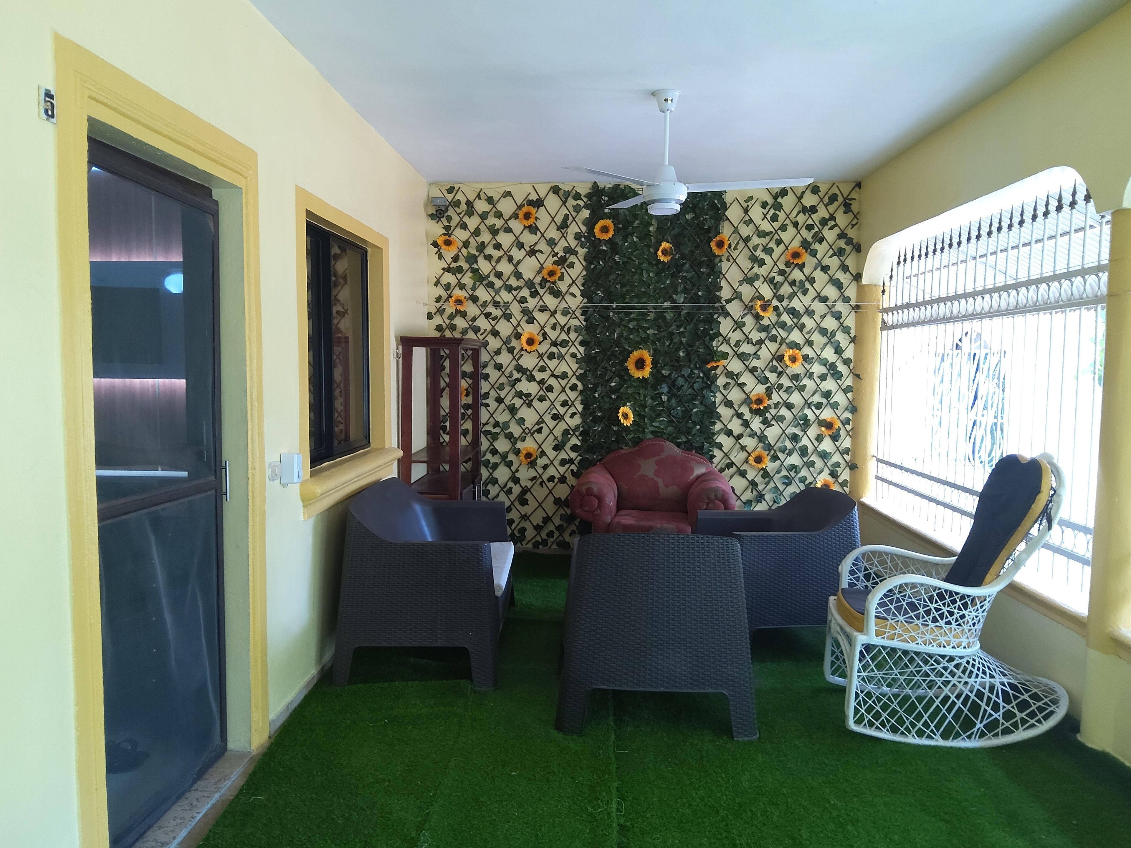 A room with a ceiling fan, a window with a metal grill, a wall with a sunflower pattern, and two wicker chairs.