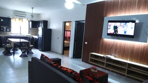 Ein modernes Wohnzimmer mit einer Couch, einem Couchtisch und einem an einer Holzwand montierten Fernseher. Im Hintergrund befindet sich ein Essbereich mit Stühlen und einem Tisch.