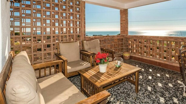 Un balcón con un separador de madera celosíaca, un sofá, dos sillas, una mesita con un florero y vistas al mar.