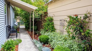 Une terrasse couverte avec des sièges d’extérieur, un jardin avec différentes plantes et fleurs, et une clôture en bois.