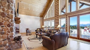 A spacious living room with a stone wall, wooden ceiling, and large windows offering a view of the outdoors. - Home on the 5th Green - Golf, Gameroom, Huge Patio (Angel Fire)