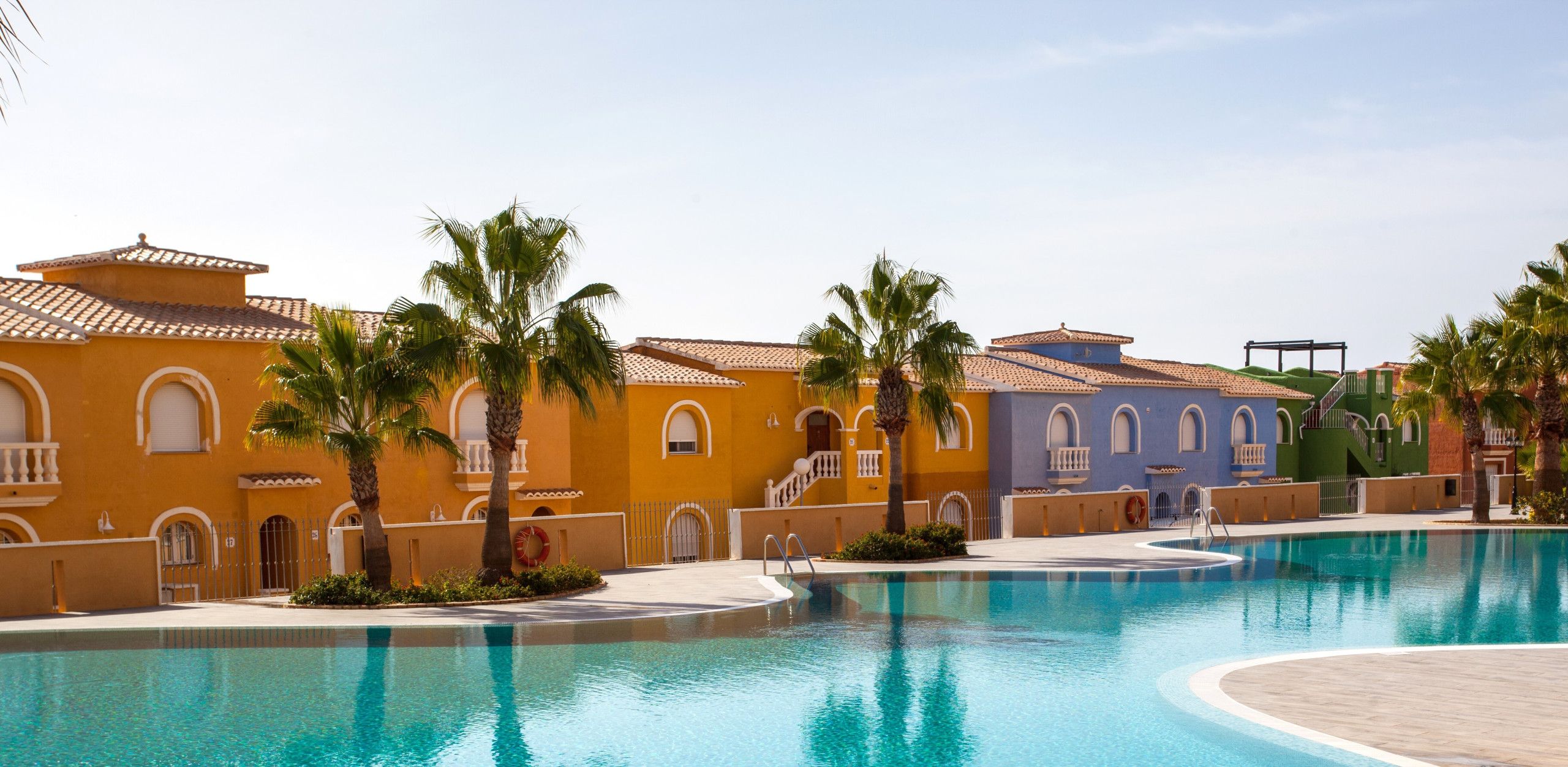 Una piscina con superficie de agua azul cristalina, rodeada de edificios de estilo arquitectónico mediterráneo, que incluyen arcos y balcones.