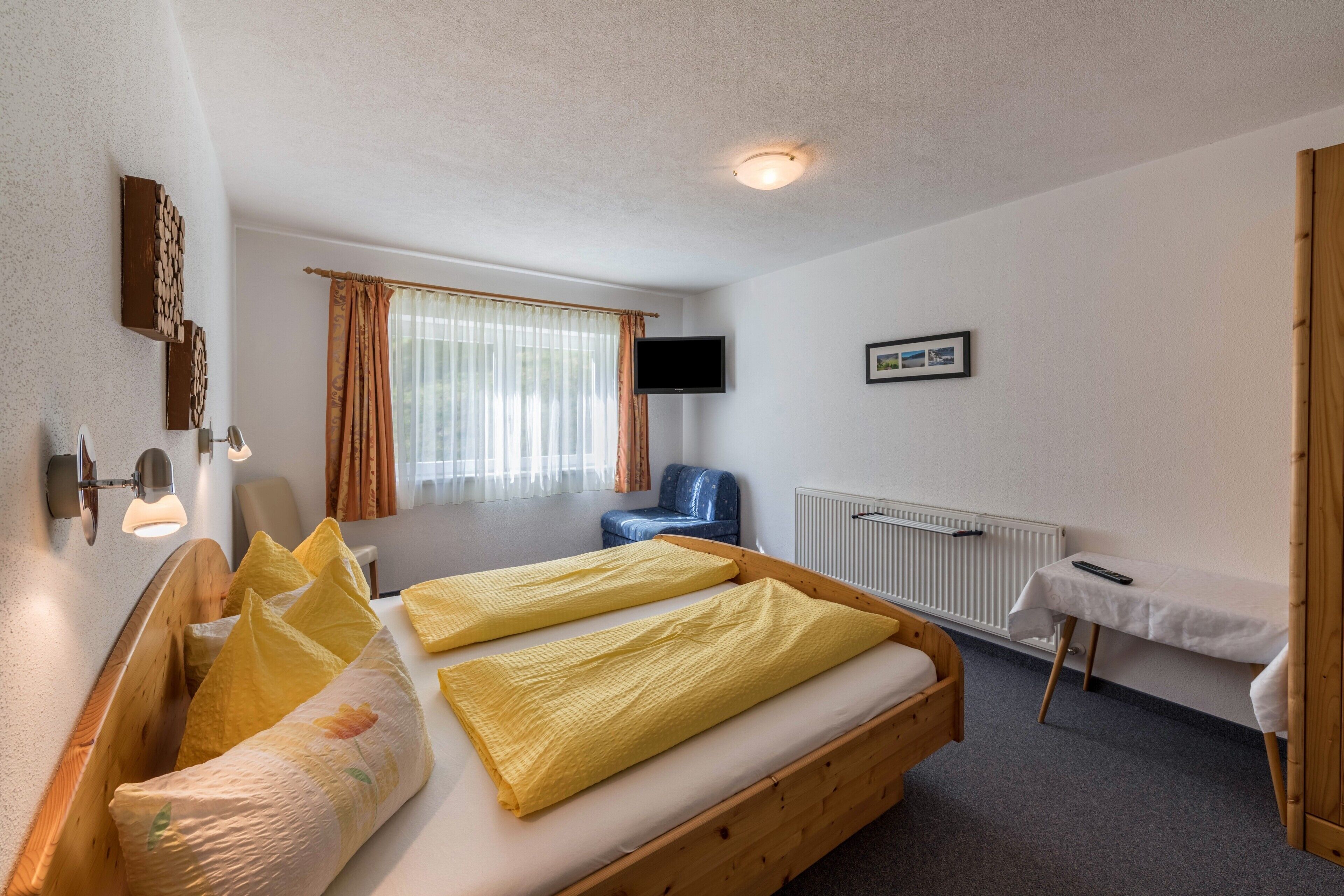 Une chambre à coucher avec un lit en bois, une chaise, une table, un téléviseur et une fenêtre avec des rideaux.