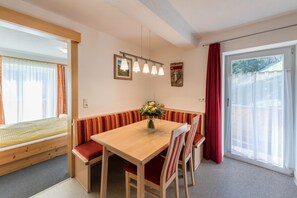 Une pièce comprenant un lit en bois, une table à manger avec des chaises, un banc, un vase avec des fleurs, un luminaire au plafond et une fenêtre avec des rideaux.