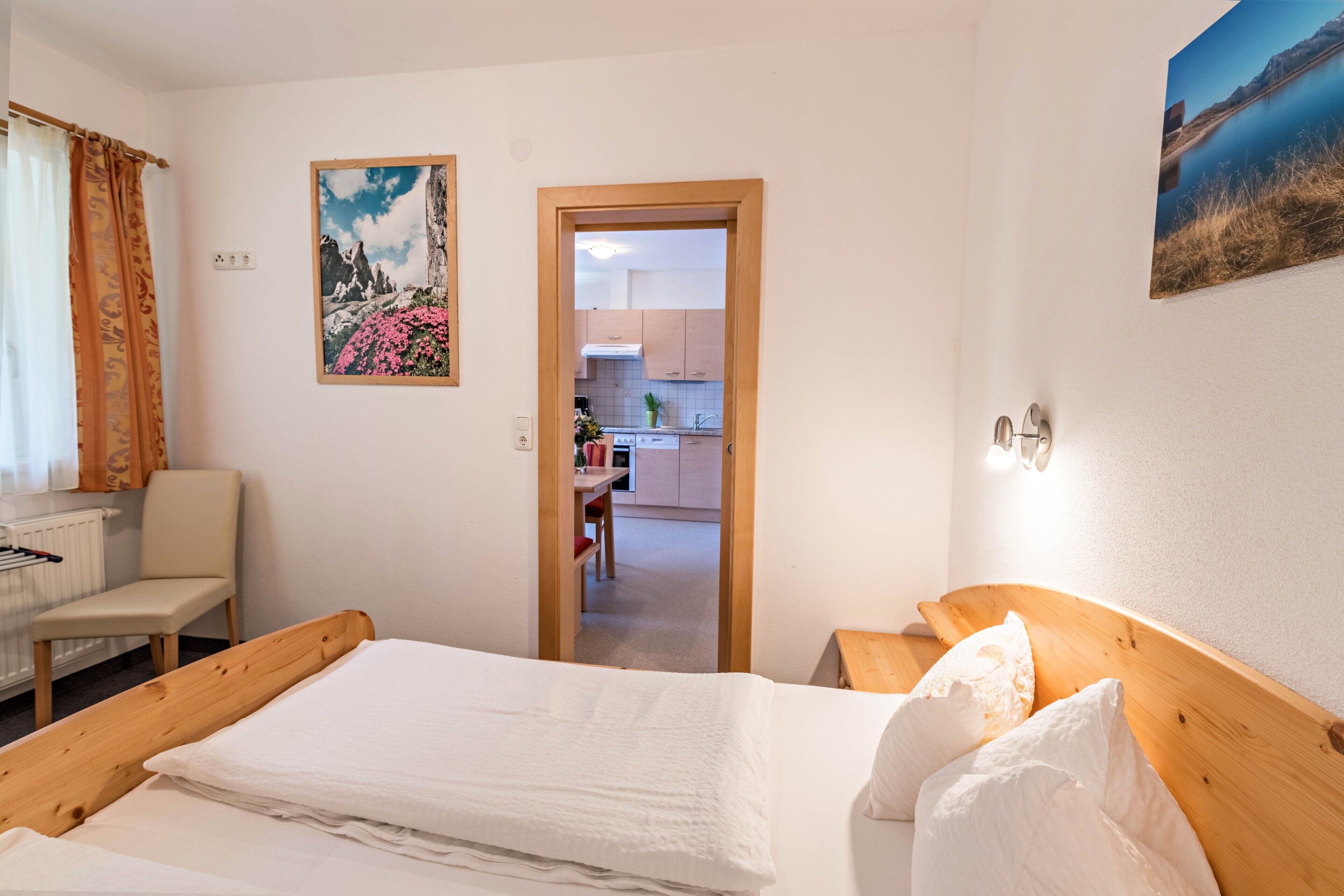 Un dormitorio con una cama de madera, una silla y vista a una cocina por una puerta abierta.