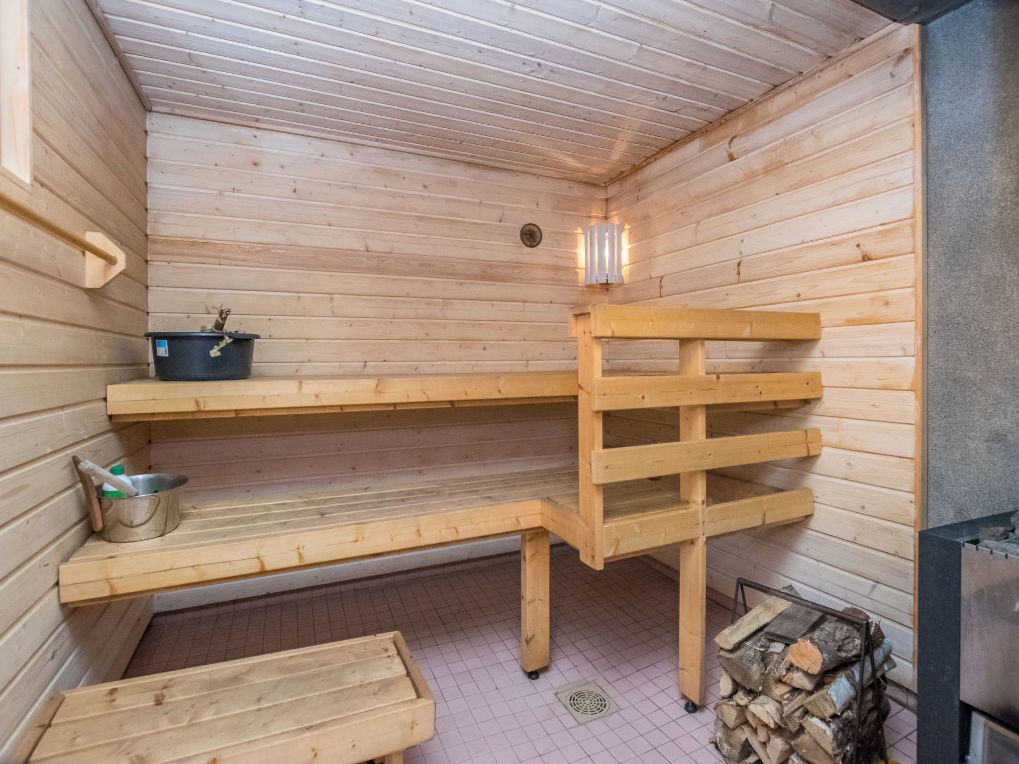 A sauna with wooden benches, a metal basin, and a stack of firewood.