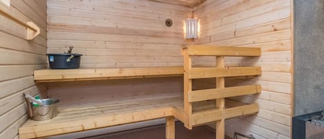 A sauna with wooden benches, a metal basin, and a stack of firewood.