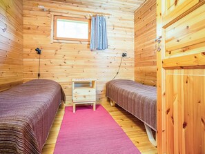 Ein Blockhauszimmer mit zwei Betten, einem kleinen Holz-Nachttisch, einem Fenster mit Vorhängen und einem rosa Teppich.