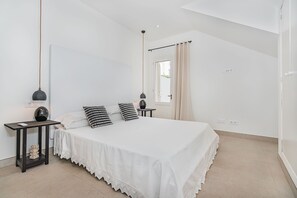 Une chambre à coucher comprenant un lit, deux tables de chevet, une commode, une lampe suspendue et une fenêtre avec des rideaux.