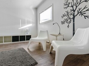 A room with two white plastic chairs, a small white table, a white bench, a grey rug, and a wall with a black tree decal. - Skivillas 78 ukkohalla (7) by Interhome (Hyrynsalmi)