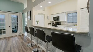 A modern kitchen with white cabinets, a stainless steel refrigerator, and a bar area with black bar stools. - Cozy Retro Chic Sarasota Home (Sarasota)