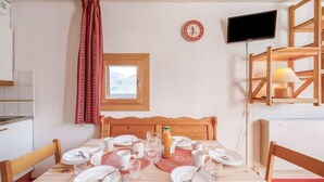 Un coin repas avec une table en bois dressée pour quatre personnes, une petite fenêtre donnant sur un paysage agréable, une horloge et un téléviseur fixé au mur.