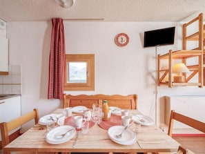 A dining area with a wooden table set for four, a small window with a view, a clock, and a TV on the wall. - Home Club (Lavachet) by Interhome (Tignes)