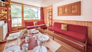 Une pièce chaleureuse avec un canapé en bois, une table à manger dressée avec des assiettes et des verres, un téléviseur posé sur un meuble et une fenêtre donnant sur un paysage verdoyant.