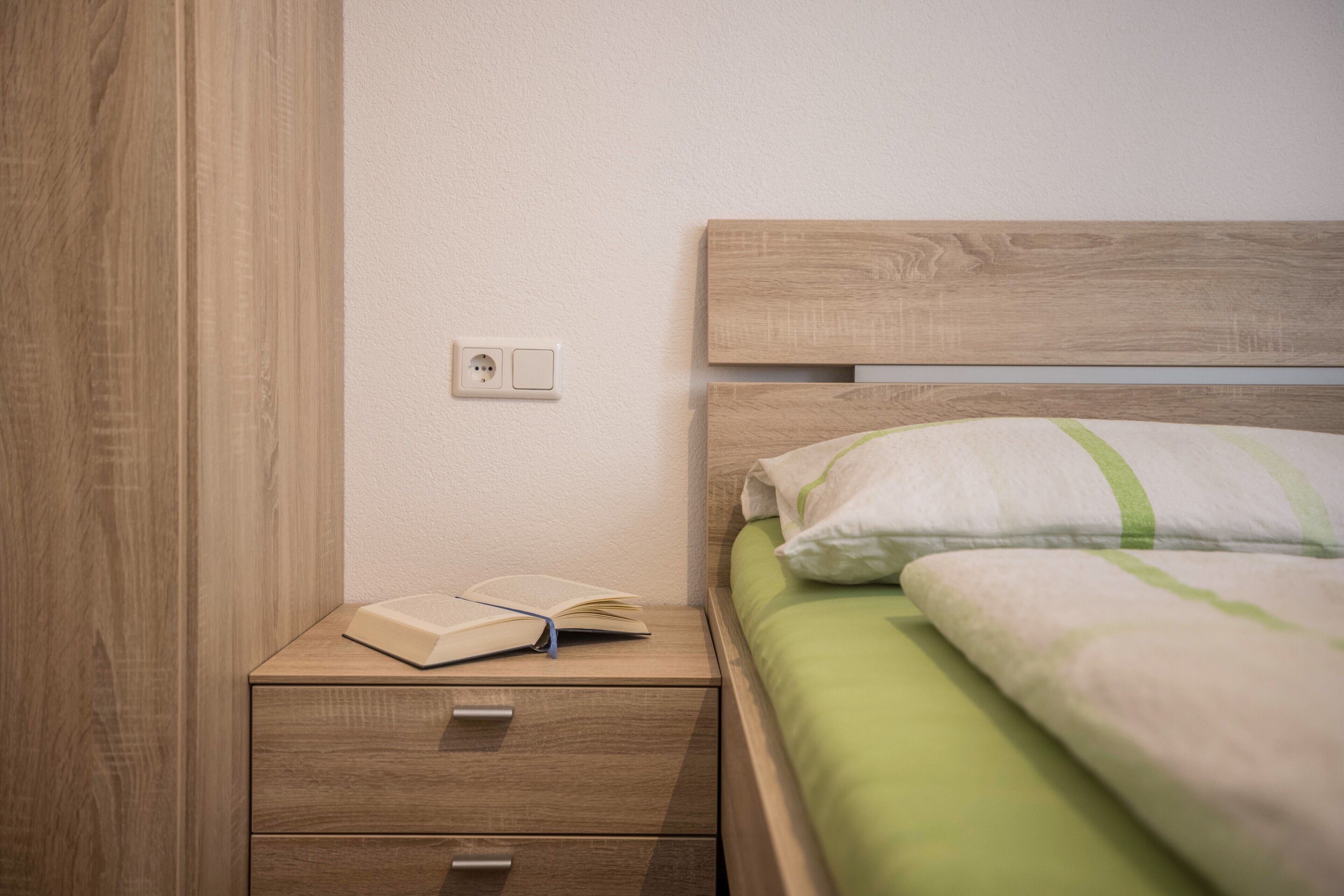 A neatly made bed with a green blanket, a wooden nightstand with a book, and a wall socket.