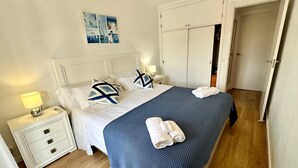 Un lit bien fait, recouvert d’une couverture bleue, avec des oreillers blancs et des serviettes enroulées, posé sur un plancher en bois.
