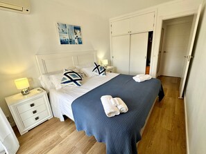 Un lit bien fait, recouvert d’une couverture bleue, avec des oreillers blancs et des serviettes enroulées, posé sur un plancher en bois.