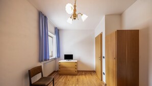 Una habitación con cama, una silla, un escritorio con computadora, armario y ventana con cortinas.