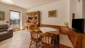 Una sala de estar con sofá, mesa de comedor de madera, sillas, televisor y una zona de cocina con armarios y ventana.