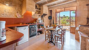 Une cuisine rustique avec un coin repas, des murs en pierre et une vue sur l’extérieur, visible à travers la porte.