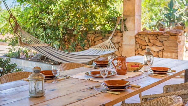 Une table en bois, dressée pour le repas, entourée de chaises en osier, avec un hamac et un mur en pierre en arrière-plan.