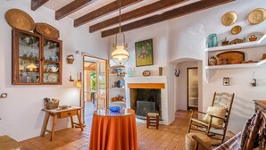Un intérieur rustique avec des poutres apparentes, une cheminée, des étagères remplies de vaisselle et un coin repas avec une table et des chaises.