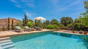Une piscine aux eaux cristallines, bordée de chaises longues et agrémentée d’un parasol, le tout entouré de verdure et d’un bâtiment.