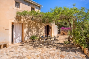 A stone courtyard with a table and chairs, a building with an arched doorway, and a tree with green foliage. - Holiday Home "Son Sunyer" with Mountain View, Pool, Garden, Balcony & Terrace (s'aranjassa)