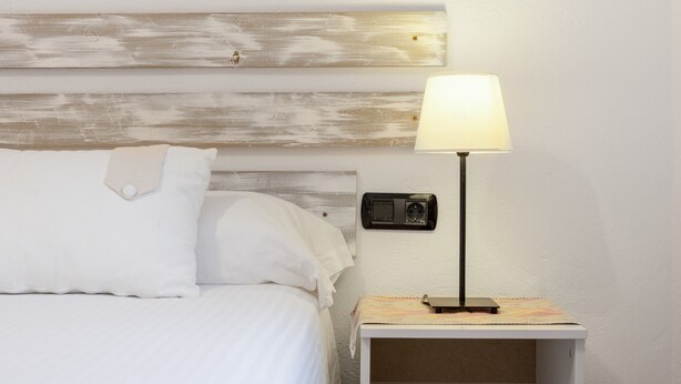A bed with white linens, a wooden headboard, a bedside lamp, and a small nightstand with a power outlet.