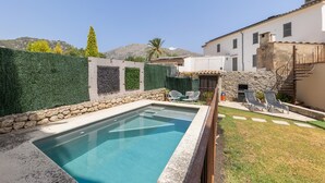 A swimming pool with a stone border, a stone wall with greenery, and a white building with a staircase.