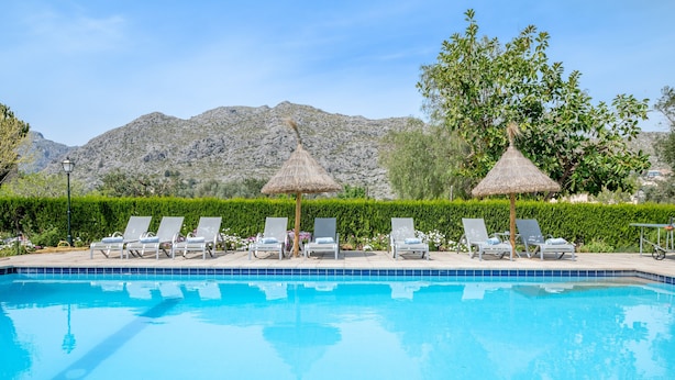 A swimming pool with lounge chairs and thatched umbrellas, surrounded by greenery and mountains. - Roomy Villa “Villa Agnes” with Mountain View, Pool, Terrace & Garden (Pollensa)