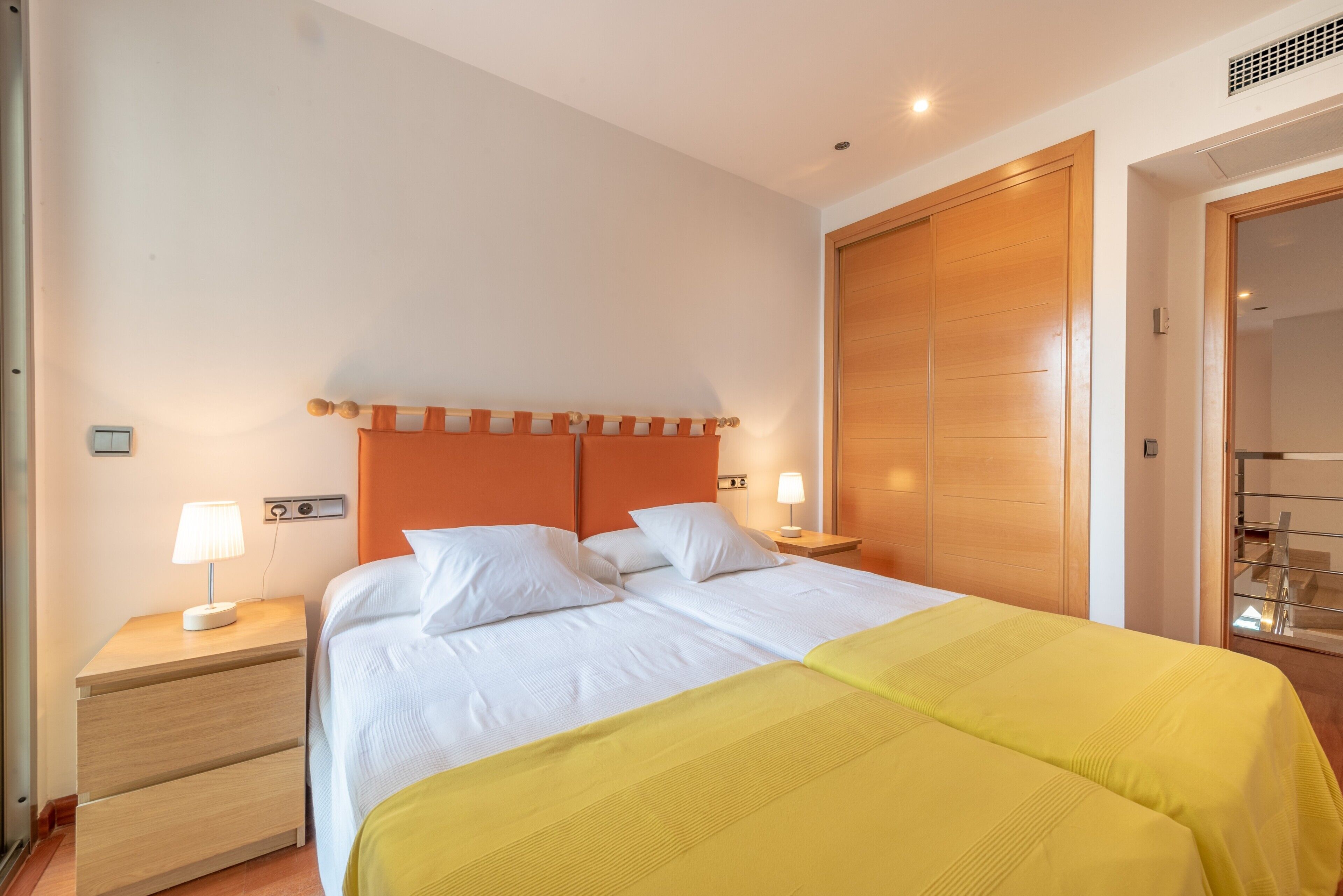 A double bed with white bedding, a wooden headboard, a bedside table with a lamp, and a wooden wardrobe.