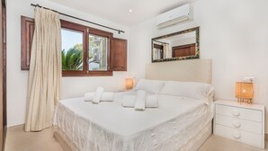 A bedroom with a bed, a window with curtains, a bedside lamp, a mirror, and a white dresser. - In perfect location to beach and city - Villa Hibiscus (Pollensa)