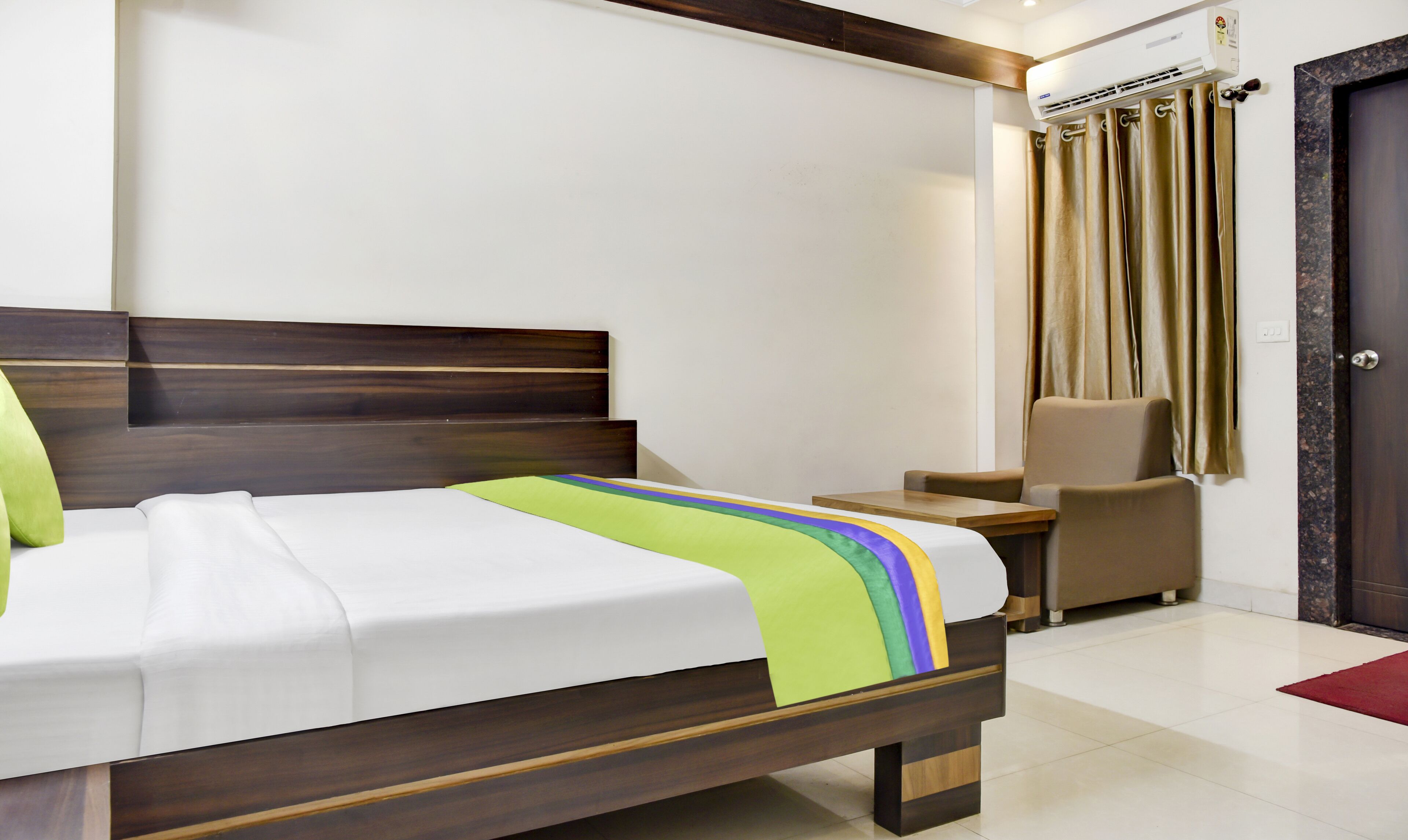 A hotel room with a bed, a small table, a chair, an air conditioning unit, and a door.