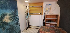 Una stanza compatta con un letto, un piccolo ripiano, una ventola e una porta con tenda.