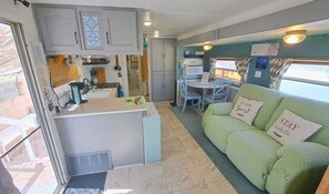 A compact RV interior with a kitchenette, a dining area, and a green couch with guest pillows. - Foothills Quiet Guest RV Home - Queen, Twin, Dogs + Fenced, BBQ, Laundry Service (Yuma)