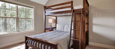 Een slaapkamer met een stapelbed, een nachtkastje met een lamp en een raam met uitzicht op bomen.