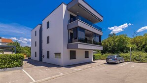 A modern two-story building with a flat roof, large windows, and a parking area in front. - Apartment LAGANINI III (Njivice)
