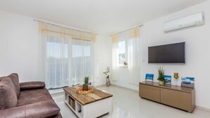A modern living room with a brown leather sofa, a wooden coffee table with a pineapple centerpiece, a white cabinet with a flat-screen TV, and a large window with sheer curtains. - Apartment LAGANINI III (Njivice)