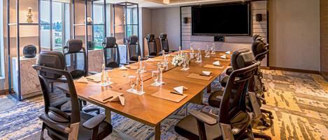 A conference room with a long table, multiple chairs, a large screen, and a city view through the windows.