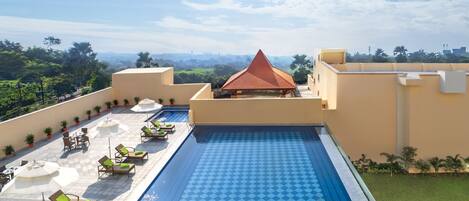 A rooftop pool with lounge chairs and umbrellas, surrounded by greenery and a clear sky.