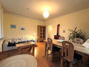 A cozy living room with a sofa, wooden dining table, chairs, and a small stool. There is a cabinet and a potted plant in the room.