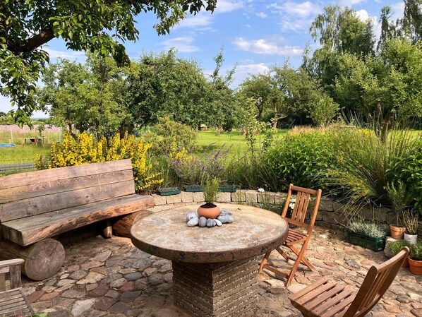 A stone table with a pot, surrounded by wooden furniture, set on a stone-paved patio with a garden view.